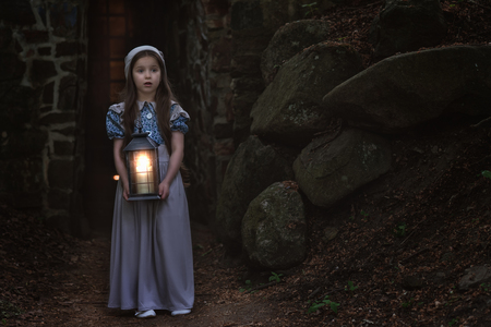 Frightened little girl in retro-style clothes is standing with a lantern near the fortress wall.の写真素材