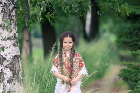 Portrait of a little beautiful girl in a birch grove. Her shoulders are covered with a shawl with traditional Russian folk patterns.の写真素材