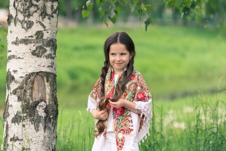 Portrait of a little beautiful girl in a birch grove. Her shoulders are covered with a shawl with...