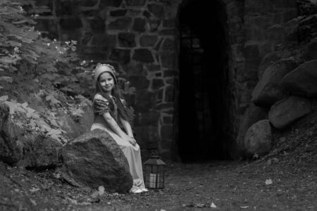 Frightened little girl in retro-style clothes is sitting with a lantern near the fortress wall. Black and white.の写真素材