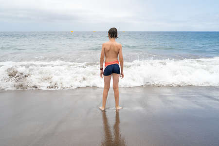 A teenager is standing on the sandy shore of the ocean.の写真素材