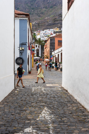GARACHICO, SPAIN - AUGUST 5, 2025: Tourists walking on cobblestone street between traditional colorful houses with mountain slope and town buildings in background. Tenerife, Canary Islands.のeditorial素材