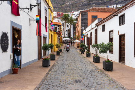 GARACHICO, SPAIN - AUGUST 5, 2025: Cobblestone street with potted trees, motorbike and walking tourists leading to historic church and palm trees. Tenerife, Canary Islands.のeditorial素材