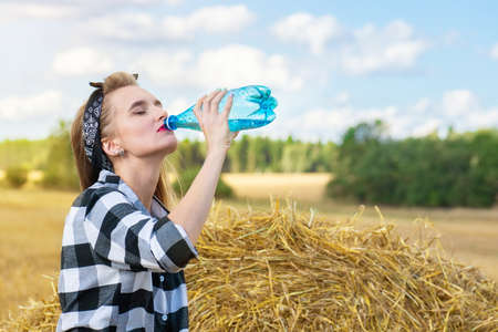 A girl is drinking water in a field near a sheaf of hay