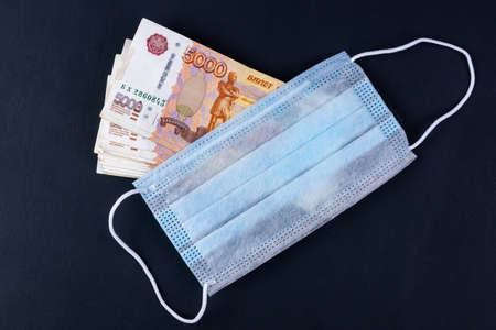 Russian money, rubles wrapped in a medical mask. Medical face masks and money. Surgical face mask. COVID-19 caused a shortage of face masks. Medical mask and five thousand rubles banknote on a black.の写真素材