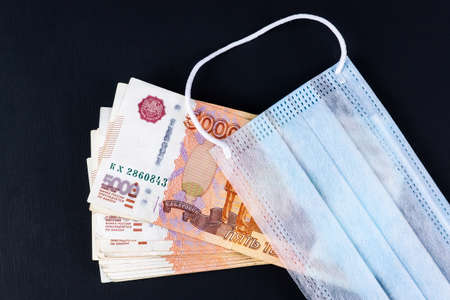 Russian money, rubles wrapped in a medical mask. Medical face masks and money. Surgical face mask. COVID-19 caused a shortage of face masks. Medical mask and five thousand rubles banknote on a black.の写真素材