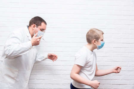 The smiling boy is looking away while his doctor holding a syringe next to his arm. Pediatrician makes vaccination to cute Caucasian boy. Boy in medical face mask getting flu shot by doctorの写真素材