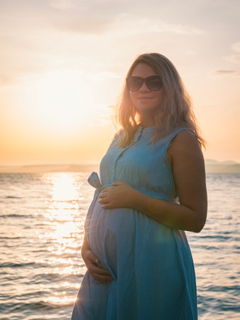 Pregnant woman in a blue dress enjoying sunset by the water at a serene locationの写真素材