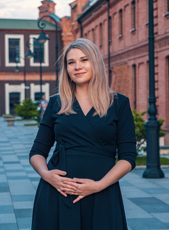 Young pregnant woman in a black dress poses confidently outside historic red brick buildings in a town square during sunsetの写真素材