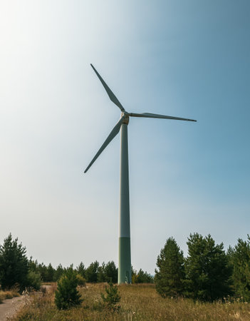 Tall wind turbine stands against a clear blue sky surrounded by green fields on a sunny day in an open landscapeの写真素材
