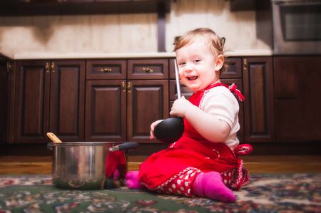 Beautiful baby in red apron with ladle in hand sitting next to a pan.の写真素材