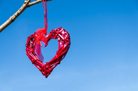 Red country wicker heart hanging from tree branch against blue sky; Valentines Day and love concept...