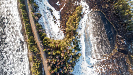 Drone view of spring rural road in yellow pine forest with melting ice lake with snow.の写真素材