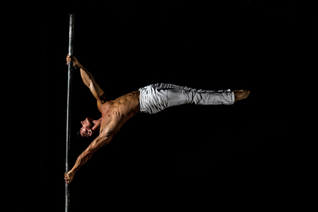 A strong muscular guy dressed in white pants performs a trick on a pole in on a black backgroundの写真素材