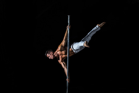 A strong muscular guy dressed in white pants performs a trick on a pole in on a black backgroundの写真素材