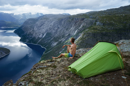 Norwegian fjord landscape with camping tent and young muscular man sitting on the edge of cliff. Norway adventureの写真素材