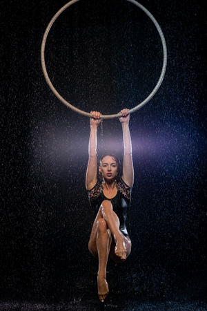 Beautiful female acrobat hanging on aerial hoop under rain on black backgroundの写真素材