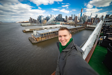 Young man makes selfie with huge cruise ship and New York city on background. Concept of happy vacation and travellingの写真素材