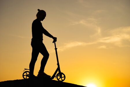 silhouette of a man standing and holding kick scooter with beautiful sunrise on backgroundの写真素材