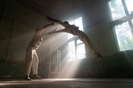 Male duo making acrobatic tricks wearing costume of insane people in abandoned room with sun rays...