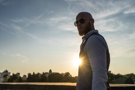 Bold Guy with a stylish beard and sunglasses on a blurred city background during sunset. Concept of success and willの写真素材