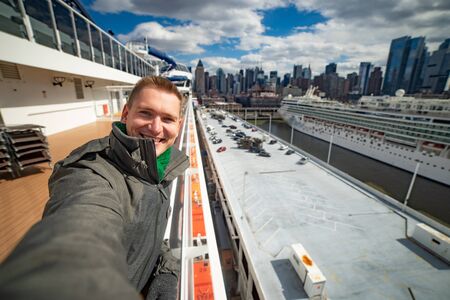 Young man makes selfie with huge cruise ship and New York city on background. Concept of happy vacation and travellingの写真素材