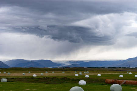 Bale of fodder grass wrapped in white plastic lying on the field during stormy cloud in Iceland.の写真素材