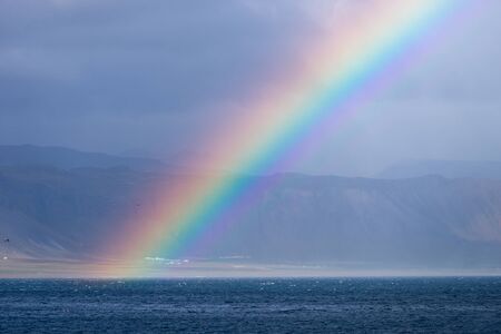 Beautiful summer landscape with sea, small village on the hill, bright colorful rainbow on blue sky, clouds and mountains on the horizon. Iceland, Peninsulaの写真素材