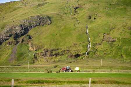 Red Tractor collecting stack of grass on green field with beautiful river going by the mountain on backgroundの写真素材