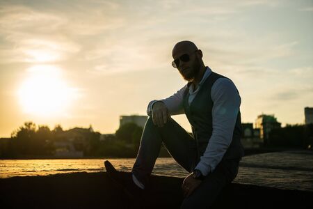 Bold Guy with a stylish beard and sunglasses on a blurred city background during dramatic sunset. Concept of success and willの写真素材