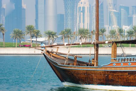 Retro historical boat with blurred panoramic view of modern skyline of Doha and green palms on background.の写真素材