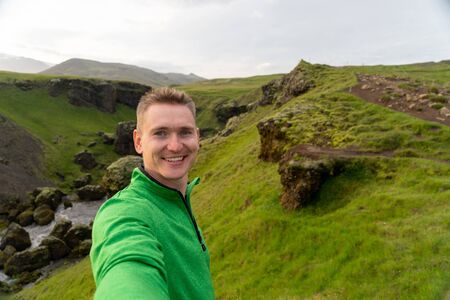 Young smiling man with river and green mountains on background takes a Selfie. Concept of freedom movement and freedomの写真素材