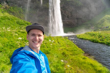 Young man taking selfie with beautiful waterfall of Seljalandsfoss on background. Concept of traveling and healthy lifestyleの写真素材