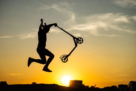 silhouette of a man riding and jumping on kick scooter with beautiful sunrise on background.の写真素材