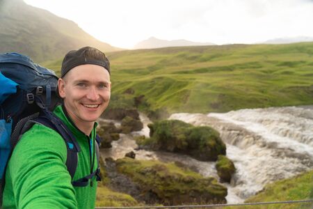 Young smiling man with river and green mountains on background takes a Selfie. Concept of freedom movement and freedomの写真素材