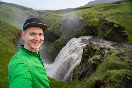 Young smiling man with waterfall and green canyon on background takes a Selfie. Concept of freedom movement and freedomの写真素材