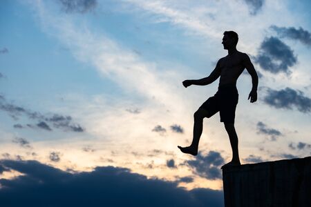 Silhouette of man against background of clouds and sunset. He stands on edge of roof. He is ready to take a step. Concept of Risk and courageの写真素材