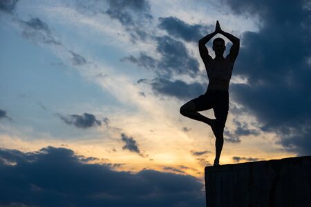 Fit young man practices sun salutation yoga on the edge of cliff at sunsetの写真素材