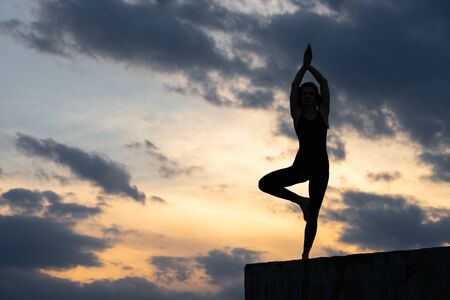 Fit young girl practices sun salutation yoga on the edge of cliff at sunsetの写真素材