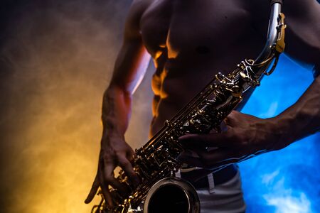 Muscular man with naked torso playing on saxophone with smoked colorful background.の写真素材