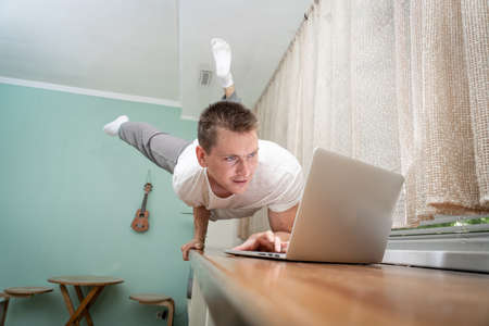 A muscular man keeps balance on one hand and works at a laptop. Self-isolation, teleworking and body training conceptの写真素材