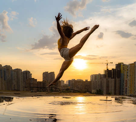 Female flexible dancer jumping during sunset on cityscape background with reflection in the water. Concept of freedom and happinessの写真素材