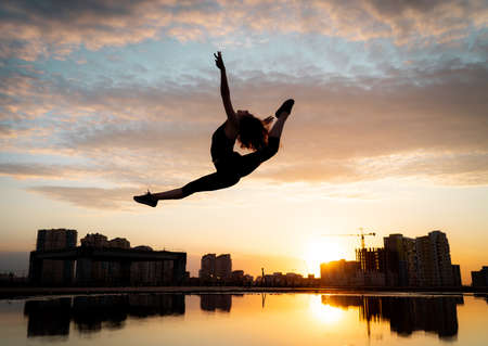 Female flexible dancer jumping during sunset on cityscape background with reflection in the water. Concept of freedom and happinessの写真素材