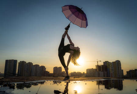 young, flexible female dancer holds an umbrella with her leg in a split on the cityscape background during sunset with reflection in the water. concept of passion and happinessの写真素材