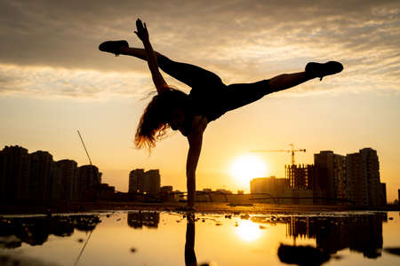 Female dancer showing her flexibility during sunset on cityscape background with reflection in the water. Concept of freedom and happinessの写真素材
