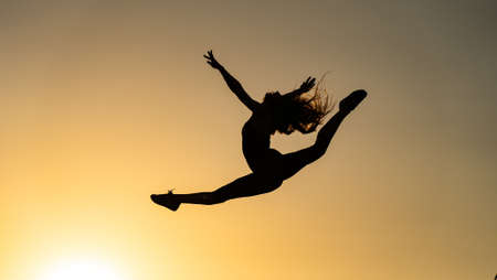 Female flexible dancer jumping during sunset on the orange sky background. Concept of freedom and happinessの写真素材