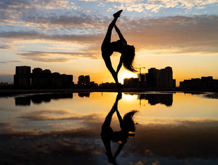 Female dancer showing her flexibility during sunset on cityscape background with reflection in the water. Concept of freedom and happinessの写真素材