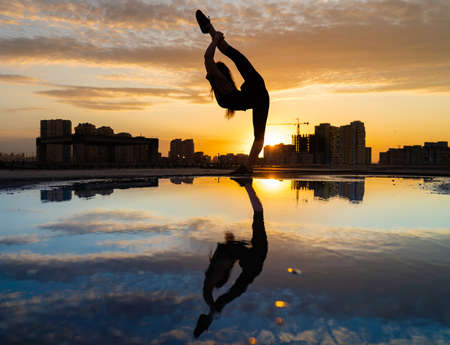 Female dancer showing her flexibility during sunset on cityscape background with reflection in the water. Concept of freedom and happinessの写真素材