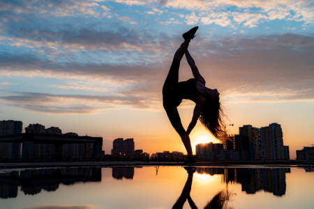 Female dancer showing her flexibility during sunset on cityscape background with reflection in the water. Concept of willpower and opportunityの写真素材
