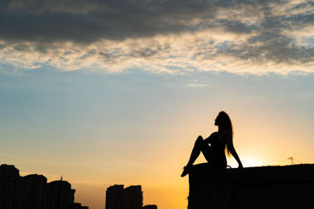 Silhouette of woman sitting on the edge of rooftop on the dramatic sky and cityscape background during sunset. Concept of challenge and choice in lifeの写真素材